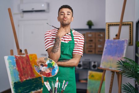 Téléchargez les photos : Brazilian young man holding painter palette at artist studio looking at the camera blowing a kiss being lovely and sexy. love expression. - en image libre de droit