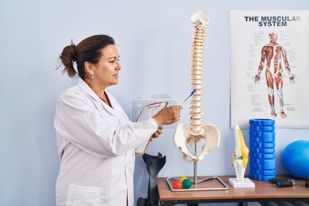 Photo for Middle age hispanic woman physiotherapist pointing to anatomical model of vertebral column at rehab clinic - Royalty Free Image