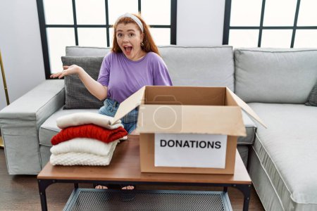 Téléchargez les photos : Young redhead woman putting used clothes into donations box celebrating achievement with happy smile and winner expression with raised hand - en image libre de droit