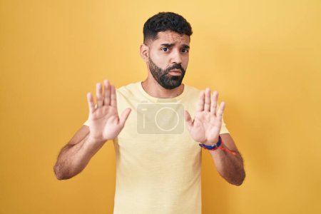 Foto de Hispanic man with beard standing over yellow background moving away hands palms showing refusal and denial with afraid and disgusting expression. stop and forbidden. - Imagen libre de derechos