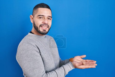 Foto de Hombre hispano de pie sobre fondo azul señalando a un lado con las manos abiertas palmeras mostrando espacio de copia, presentando anuncio sonriendo emocionado feliz - Imagen libre de derechos