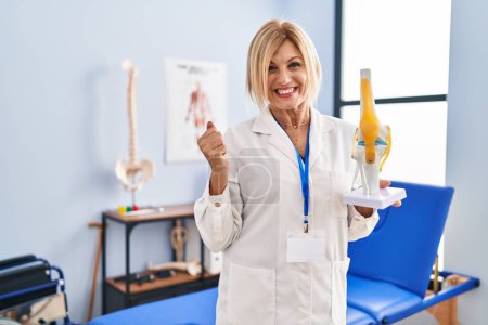 Foto de Mujer traumatóloga rubia de mediana edad sosteniendo modelo anatómico de articulación de rodilla en la clínica gritando orgulloso, celebrando la victoria y el éxito muy emocionado con los brazos levantados - Imagen libre de derechos