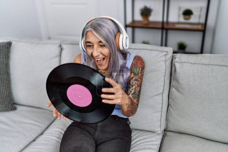 Photo for Middle age grey-haired woman listening to music holding vinyl disc at home smiling and laughing hard out loud because funny crazy joke. - Royalty Free Image