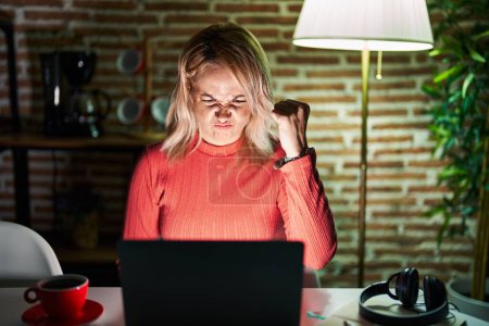 Foto de Mujer rubia usando el ordenador portátil por la noche en casa enojado y loco levantando el puño frustrado y furioso mientras gritaba con ira. rabia y concepto agresivo. - Imagen libre de derechos