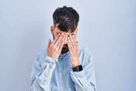 Téléchargez les photos : Young hispanic man with beard standing over blue background rubbing eyes for fatigue and headache, sleepy and tired expression. vision problem - en image libre de droit