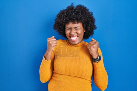 Téléchargez les photos : Femme noire avec les cheveux bouclés debout sur fond bleu excité pour le succès avec les bras levés et les yeux fermés célébrant la victoire en souriant. concept gagnant. - en image libre de droit