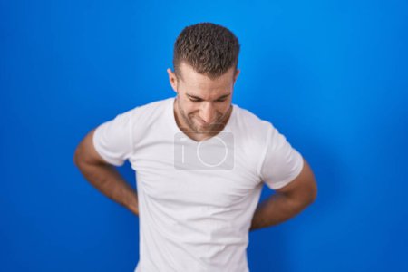 Foto de Joven hombre caucásico de pie sobre fondo azul sufriendo de dolor de espalda, tocando la espalda con la mano, dolor muscular - Imagen libre de derechos