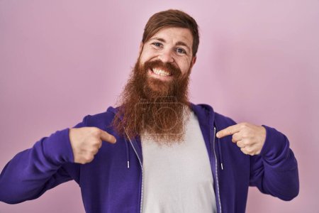 Foto de Hombre caucásico con barba larga de pie sobre fondo rosa mirando confiado con sonrisa en la cara, señalándose con los dedos orgullosos y felices. - Imagen libre de derechos