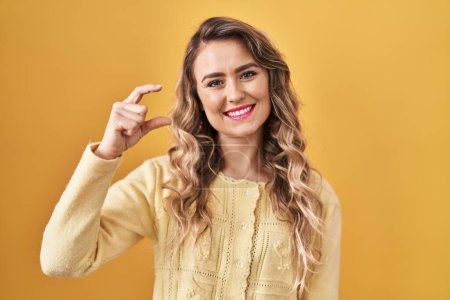 Téléchargez les photos : Jeune femme caucasienne debout sur fond jaune geste souriant et confiant avec la main faisant signe de petite taille avec les doigts regardant et la caméra. concept de mesure. - en image libre de droit