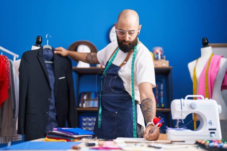 Photo for Young bald man tailor holding jacket writing on notebook at clothing factory - Royalty Free Image