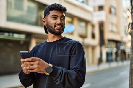 Téléchargez les photos : Jeune homme arabe souriant confiant en utilisant smartphone à la rue - en image libre de droit