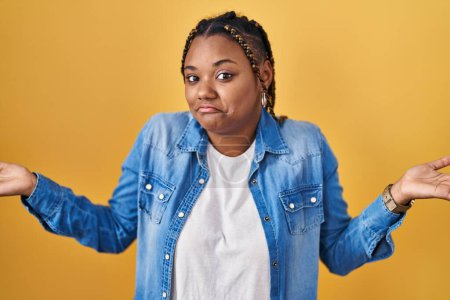 Photo for African american woman with braids standing over yellow background clueless and confused expression with arms and hands raised. doubt concept. - Royalty Free Image
