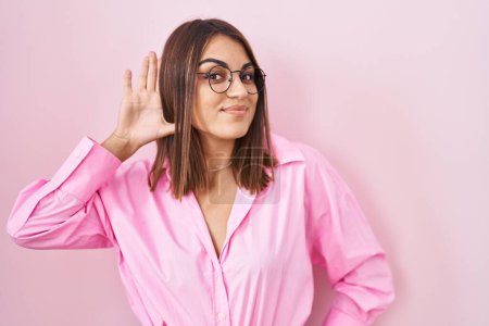 Téléchargez les photos : Young hispanic woman wearing glasses standing over pink background smiling with hand over ear listening an hearing to rumor or gossip. deafness concept. - en image libre de droit