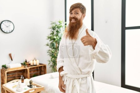 Téléchargez les photos : Homme rousse à longue barbe portant un peignoir au spa de bien-être souriant heureux et positif, pouce levé faire excellent et signe d'approbation - en image libre de droit