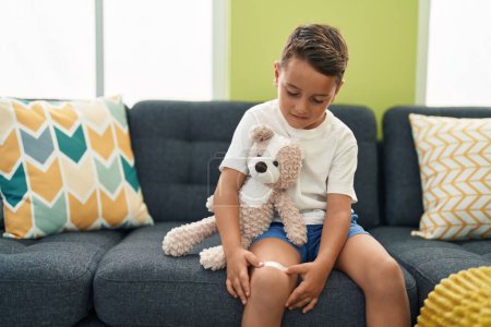 Photo for Adorable hispanic toddler hoding teddy bear putting band aid on leg at home - Royalty Free Image