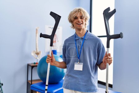 Téléchargez les photos : Jeune homme blond pysiothérapeute souriant confiant tenant des béquilles à la clinique de réadaptation - en image libre de droit