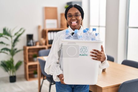 Photo for African american woman working at the office holding plastic bottle for recycling sticking tongue out happy with funny expression. - Royalty Free Image