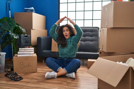 Photo for Young hispanic woman sitting on the floor at new home in shock face, looking skeptical and sarcastic, surprised with open mouth - Royalty Free Image