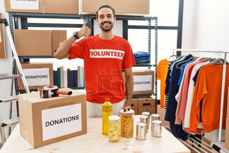 Foto de Young hispanic man wearing volunteer t shirt at donations stand doing happy thumbs up gesture with hand. approving expression looking at the camera showing success. - Imagen libre de derechos