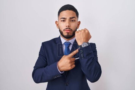 Téléchargez les photos : Young hispanic man wearing business suit and tie in hurry pointing to watch time, impatience, looking at the camera with relaxed expression - en image libre de droit