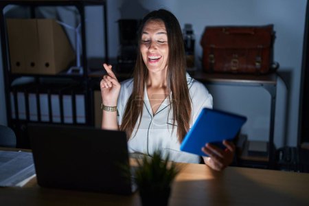 Téléchargez les photos : Young brunette woman working at the office at night gesturing finger crossed smiling with hope and eyes closed. luck and superstitious concept. - en image libre de droit