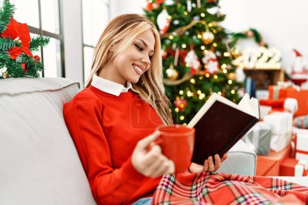 Téléchargez les photos : Jeune femme caucasienne lecture livre et boire du café assis sur le canapé par arbre de Noël à la maison - en image libre de droit