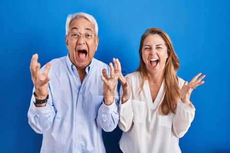 Photo for Middle age hispanic couple standing over blue background crazy and mad shouting and yelling with aggressive expression and arms raised. frustration concept. - Royalty Free Image