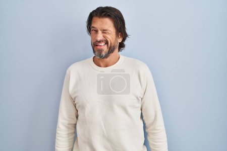 Foto de Hombre guapo de mediana edad con suéter casual sobre fondo azul guiño mirando a la cámara con expresión sexy, cara alegre y feliz. - Imagen libre de derechos