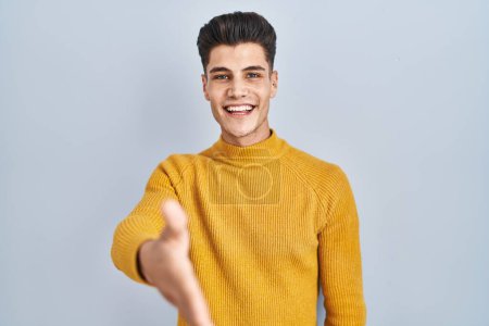 Foto de Joven hombre hispano de pie sobre fondo azul sonriente amistoso ofreciendo apretón de manos como saludo y bienvenida. negocios exitosos. - Imagen libre de derechos
