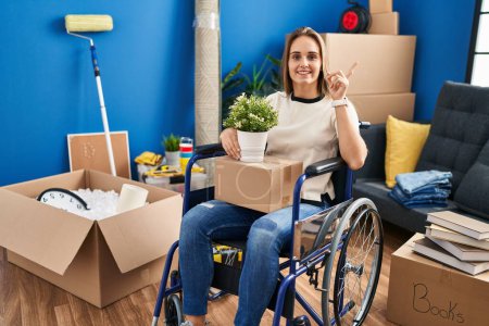 Photo for Young woman sitting on wheelchair moving to a new home with a big smile on face, pointing with hand finger to the side looking at the camera. - Royalty Free Image