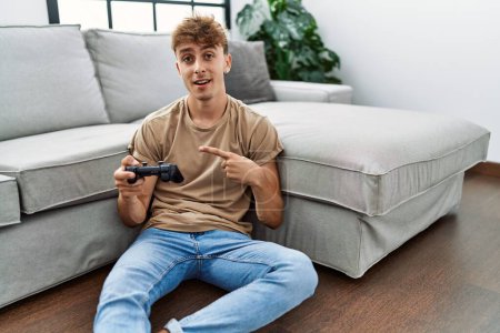 Téléchargez les photos : Young caucasian man playing video game holding controller at home smiling happy pointing with hand and finger - en image libre de droit