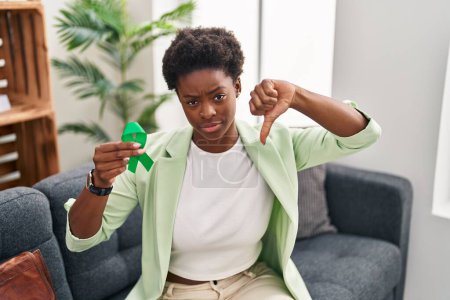 Foto de Mujer afroamericana sosteniendo apoyo cinta verde con la cara enojada, signo negativo que muestra aversión con los pulgares hacia abajo, concepto de rechazo - Imagen libre de derechos