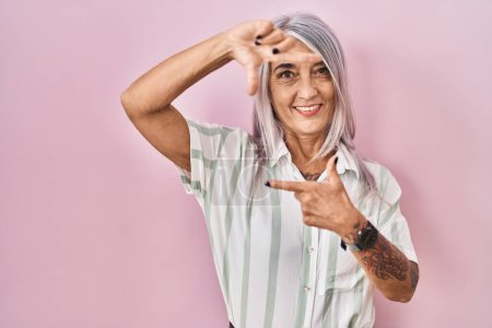 Téléchargez les photos : Femme d'âge moyen avec les cheveux gris debout sur fond rose souriant faisant cadre avec les mains et les doigts avec le visage heureux. concept de créativité et photographie. - en image libre de droit