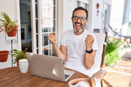 Foto de Hombre de mediana edad utilizando ordenador portátil en casa muy feliz y emocionado haciendo gesto ganador con los brazos levantados, sonriendo y gritando por el éxito. concepto de celebración. - Imagen libre de derechos