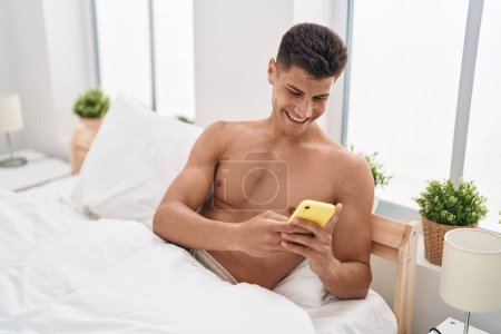 Téléchargez les photos : Young hispanic man using smartphone sitting on bed at bedroom - en image libre de droit