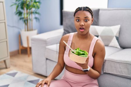 Foto de African american woman with braids eating salad after working out at home scared and amazed with open mouth for surprise, disbelief face - Imagen libre de derechos