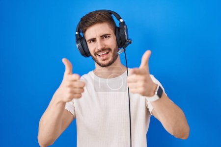Foto de Hombre hispano con barba escuchando música usando auriculares apuntando con los dedos a la cámara con cara alegre y divertida. buena energía y vibraciones. - Imagen libre de derechos