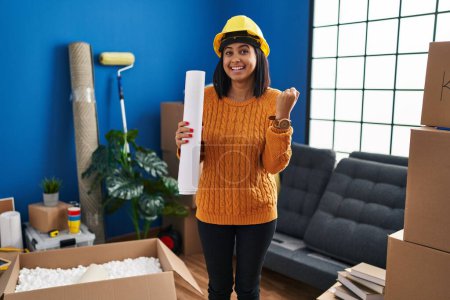 Photo for Young hispanic woman standing on new home with blueprints screaming proud, celebrating victory and success very excited with raised arms - Royalty Free Image