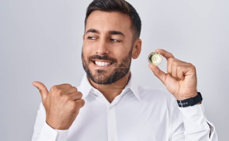 Téléchargez les photos : Bel homme hispanique tenant cardano pièce de crypto-monnaie pointant pouce vers le côté souriant heureux avec bouche ouverte - en image libre de droit
