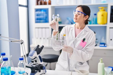 Téléchargez les photos : Jeune femme portant l'uniforme scientifique tenant des éprouvettes avec des fleurs au laboratoire - en image libre de droit
