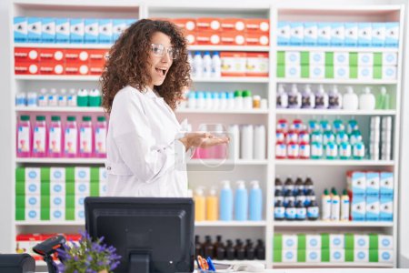 Foto de Hispanic woman with curly hair working at pharmacy drugstore pointing aside with hands open palms showing copy space, presenting advertisement smiling excited happy - Imagen libre de derechos
