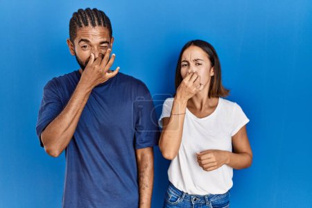 Photo for Young hispanic couple standing together smelling something stinky and disgusting, intolerable smell, holding breath with fingers on nose. bad smell - Royalty Free Image