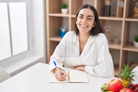 Téléchargez les photos : Jeune femme hispanique écrivant sur un cahier assis sur une table à la maison - en image libre de droit