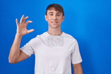 Téléchargez les photos : Caucasian blond man standing over blue background smiling and confident gesturing with hand doing small size sign with fingers looking and the camera. measure concept. - en image libre de droit
