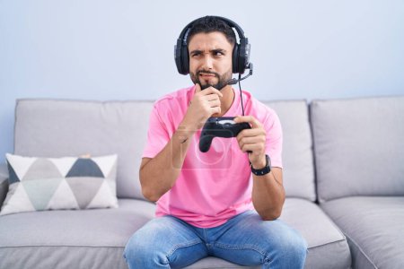 Téléchargez les photos : Jeune homme hispanique jouant contrôleur de tenue de jeu vidéo assis sur le canapé pensant inquiet d'une question, préoccupé et nerveux avec la main sur le menton - en image libre de droit