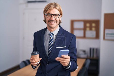 Foto de Joven hombre rubio trabajador de negocios usando teléfono inteligente beber café en la oficina - Imagen libre de derechos