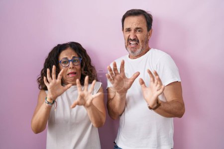 Photo for Middle age hispanic couple together over pink background afraid and terrified with fear expression stop gesture with hands, shouting in shock. panic concept. - Royalty Free Image