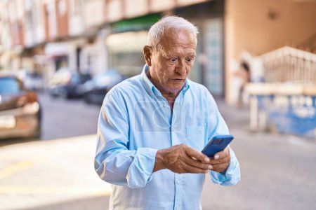 Téléchargez les photos : Homme âgé aux cheveux gris utilisant un smartphone avec une expression surprise dans la rue - en image libre de droit
