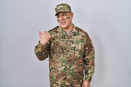 Téléchargez les photos : Jeune homme hispanique portant uniforme de camouflage de l'armée souriant avec le visage heureux regardant et pointant vers le côté avec le pouce vers le haut. - en image libre de droit