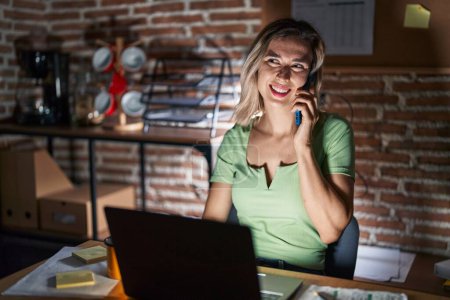 Foto de Joven hermosa mujer trabajando en la oficina por la noche hablando por teléfono sonriendo mirando a un lado y mirando hacia otro pensando. - Imagen libre de derechos